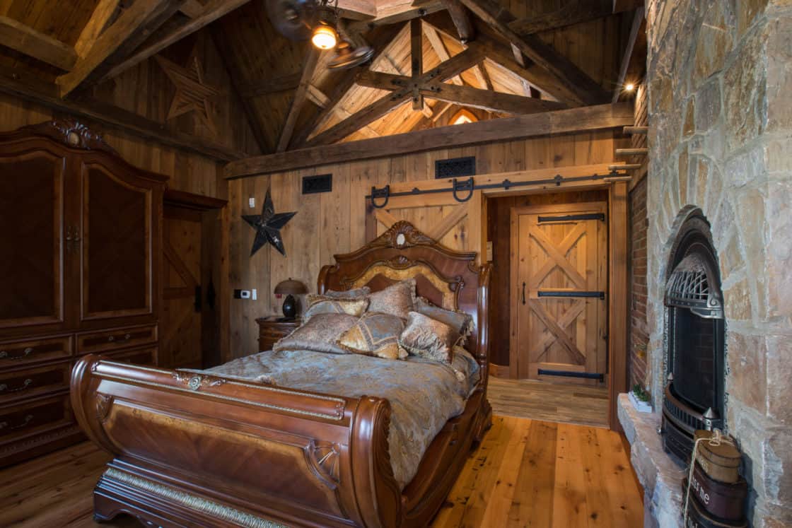 Rustic bedroom with exposed wooden beams, wooden walls, a decorative wooden bed with plush bedding, a stone fireplace, barn-style doors inspired by a party barn, and a wooden armoire and dresser. Warm, cozy atmosphere.