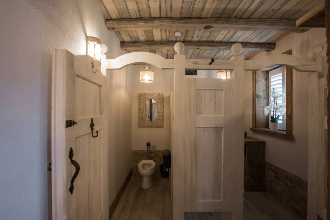 A rustic bathroom in a party barn, featuring wooden stalls, light wood paneling, exposed beams, a toilet, mirror, and a window with a potted plant. Warm lighting enhances the cozy atmosphere.