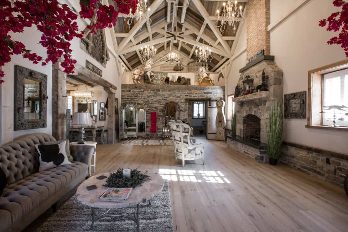 A spacious, rustic living room with exposed beams, a stone fireplace, chandeliers, and tufted sofa evokes the charm of a luxury horse barn. Light wood floors add warmth; large windows and red flowers hanging above complete the inviting ambiance.