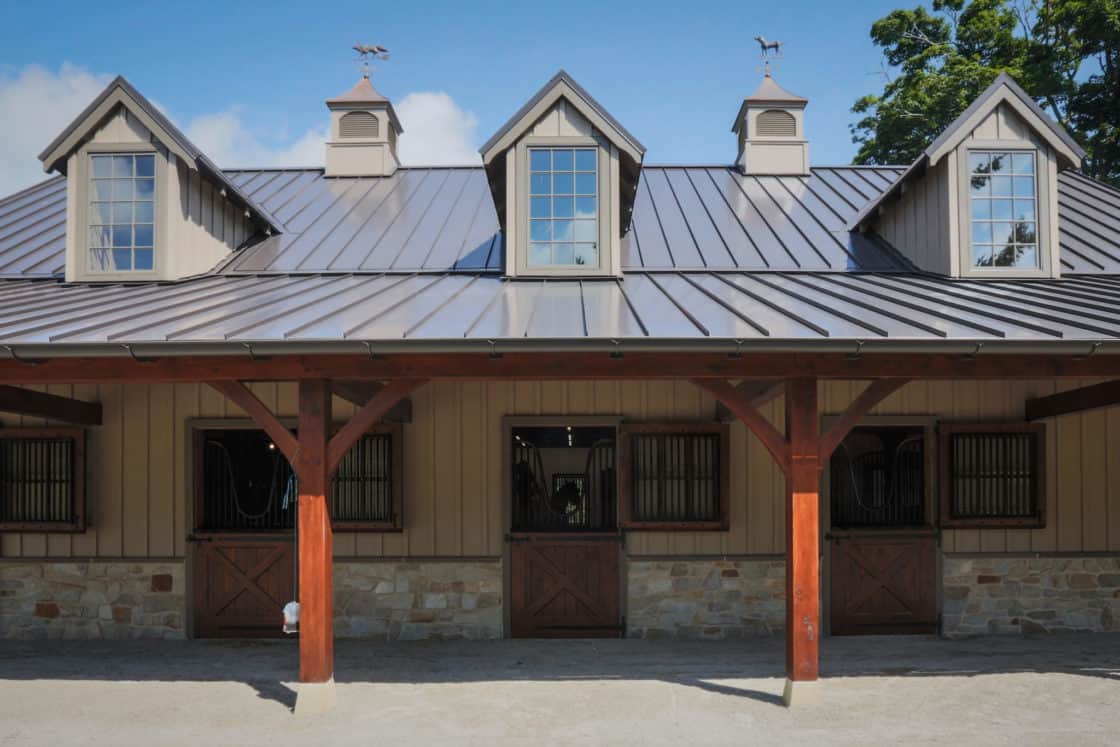 A stable party barn with a metal roof, three dormer windows, and wooden doors below. The exterior features wooden beams, stone accents, and cupolas with weather vanes on top against a blue sky.