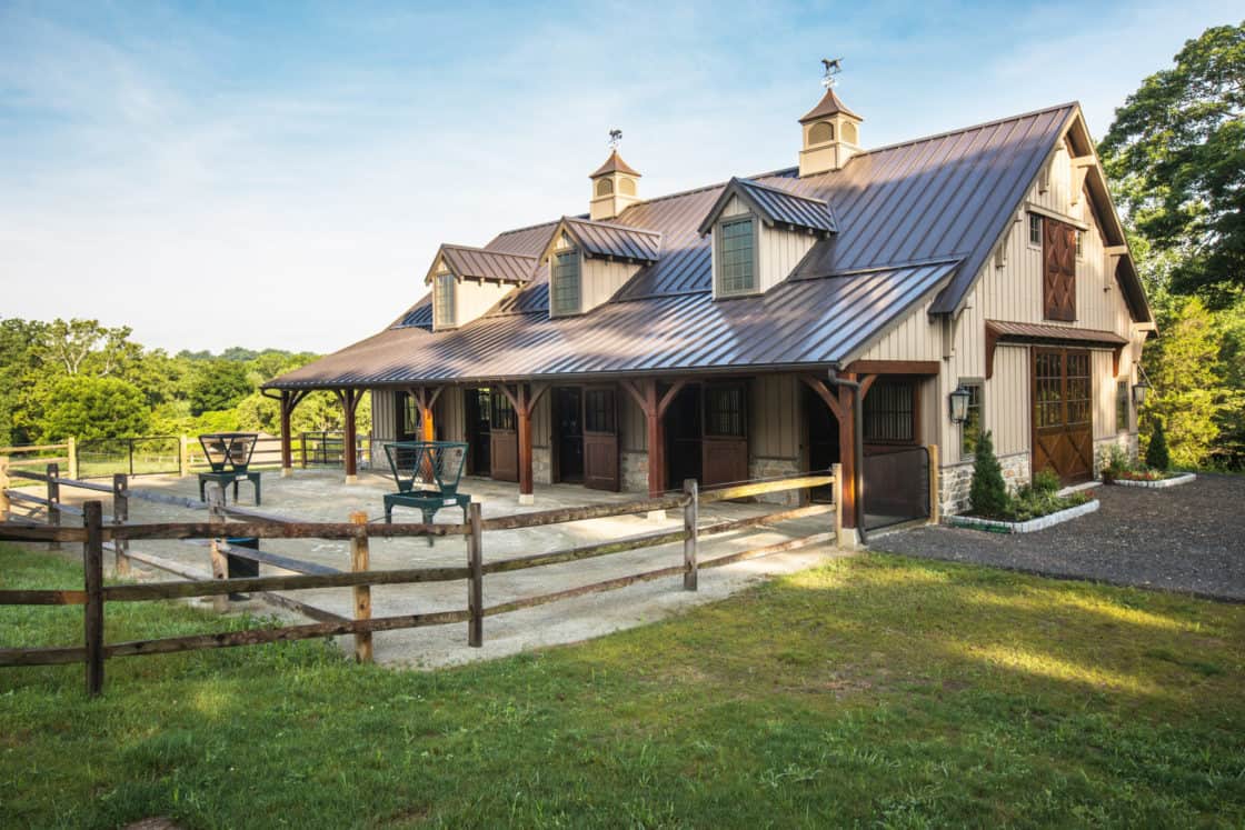 A luxury horse barn with a metal roof, cupolas, and wooden doors sits in a fenced green area with picnic tables and trees in the background under a blue sky.
