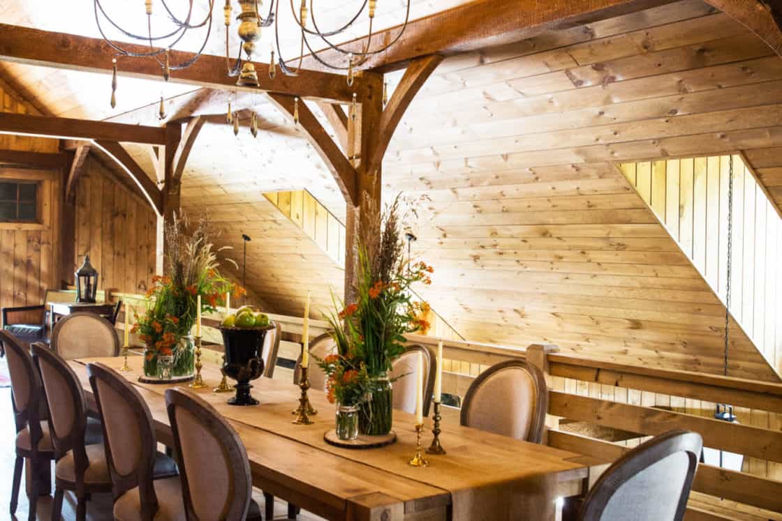 A rustic dining room in a luxury home features a long wooden table, elegant upholstered chairs, floral centerpieces, and a gold chandelier, all set in a wood-paneled room with sloped ceilings and large windows reminiscent of a classic bank barn.