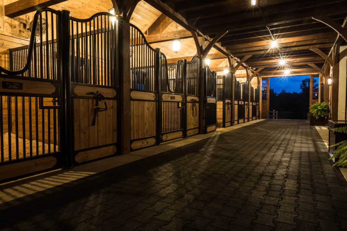 A well-lit, tidy luxury horse barn at dusk features wooden stalls along a paved aisle, iron gates, and overhead lights that illuminate the elegant space.