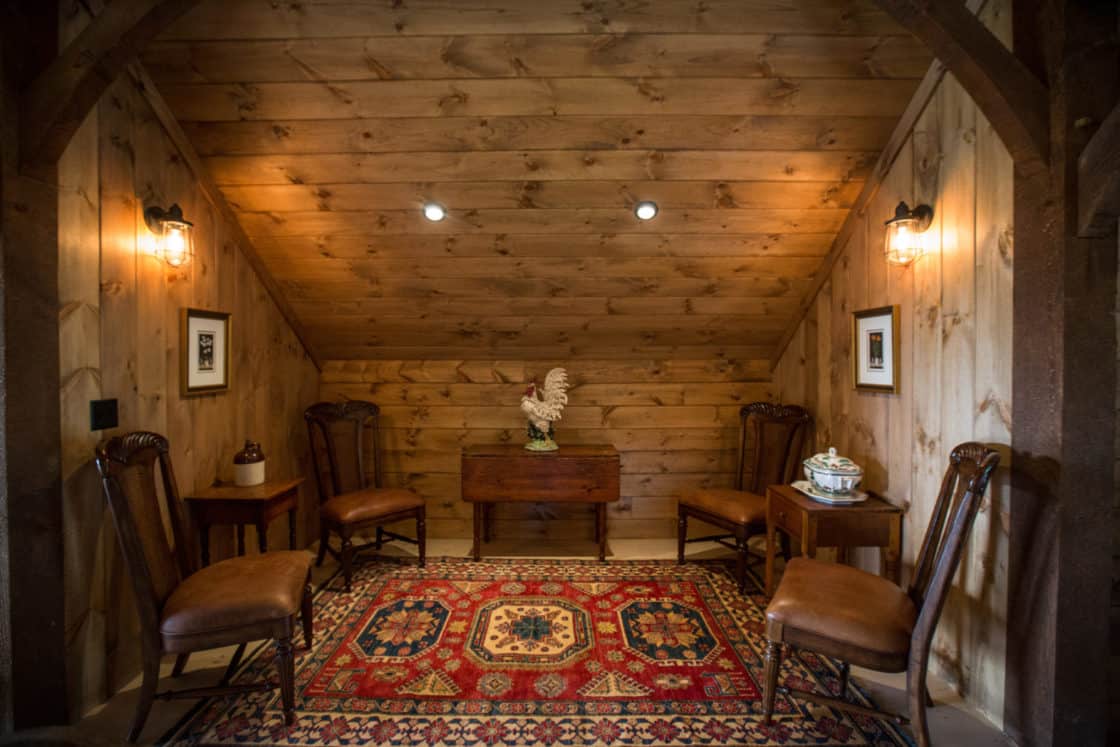 A cozy, wood-paneled sitting area in this luxury horse barn features four chairs, a small table with a decorative rooster, framed art on the walls, and a patterned red rug on the floor under warm lighting.