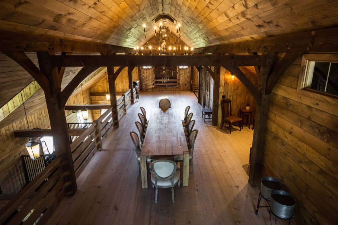 A rustic, wooden loft with exposed beams features a long dining table surrounded by chairs, a chandelier overhead, and warm ambient lighting—perfect for gatherings in this stylish party barn with a cozy, inviting atmosphere.