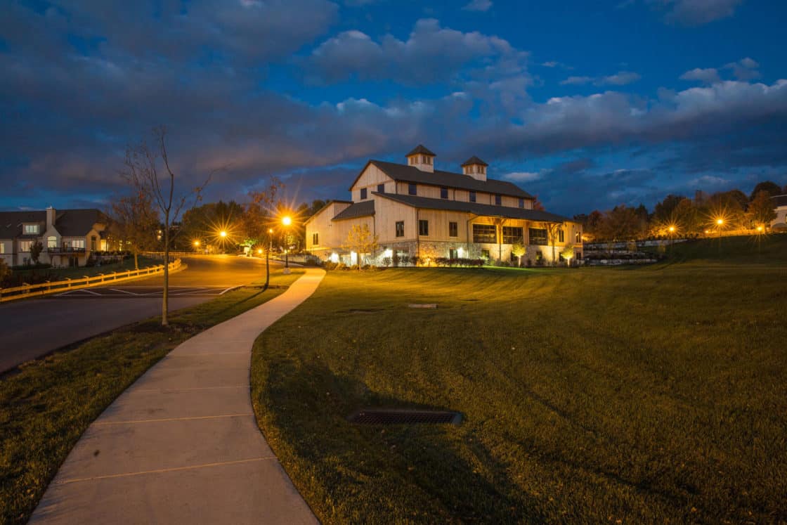 A large, barn-style building—reminiscent of a party barn—is illuminated at dusk by glowing streetlights. The sky is partly cloudy, and a sidewalk curves through the grassy lawn. Trees and houses are visible in the background.