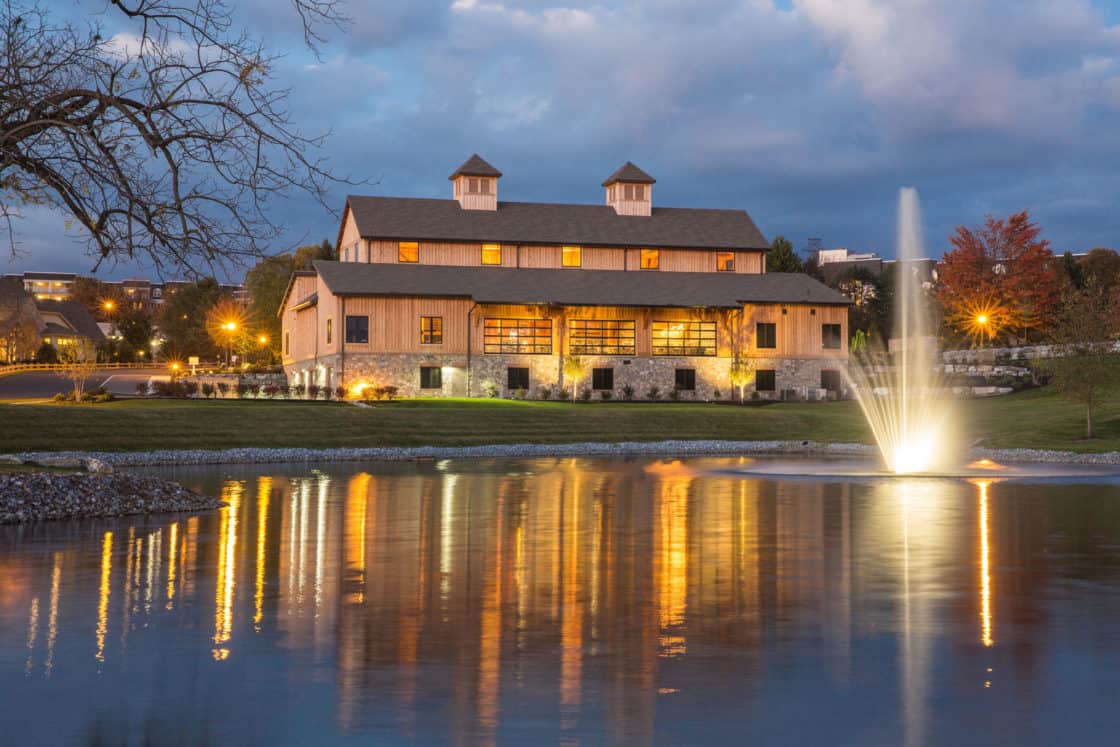 A rustic party barn with two cupolas is lit warmly at dusk, reflected in a pond with a fountain spraying water, surrounded by trees and landscaping under a cloudy sky.