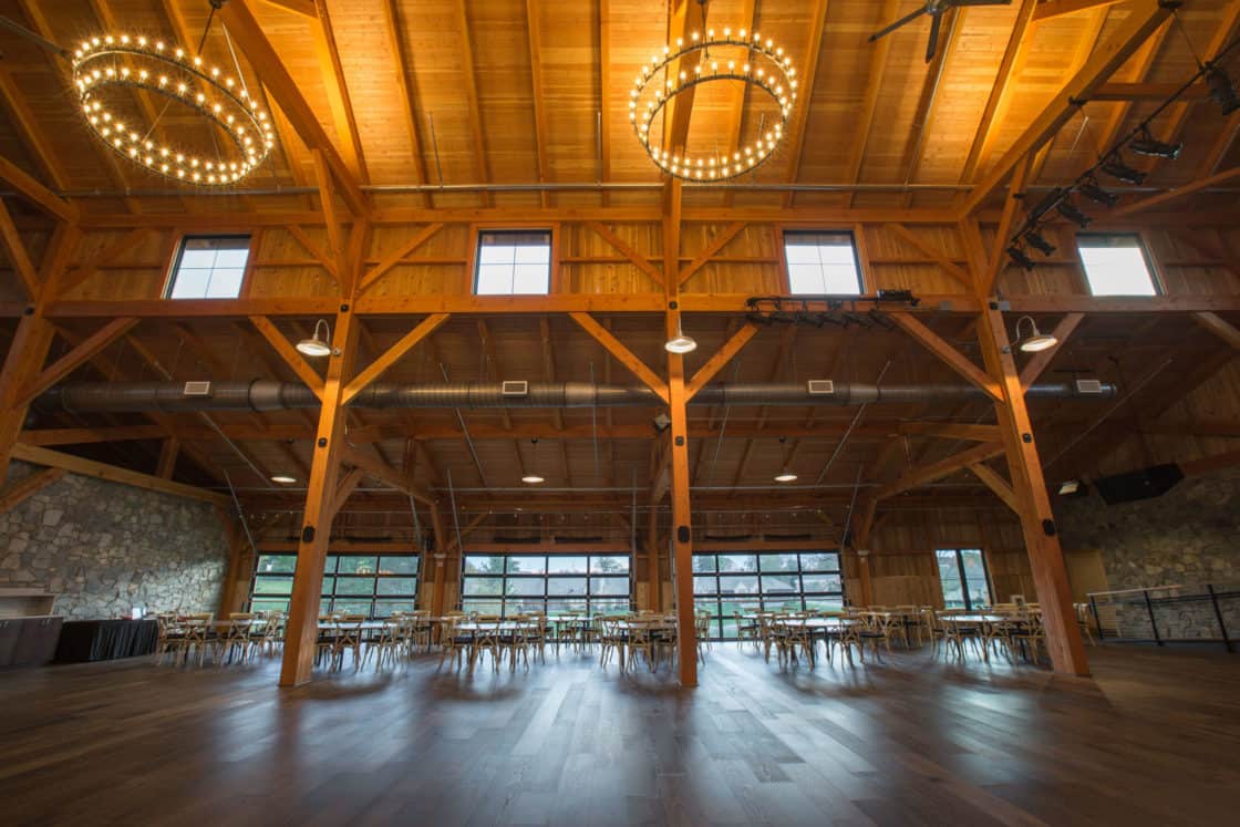 A spacious wooden hall reminiscent of a luxury horse barn, with exposed beams, stone walls, and large windows. Circular chandeliers hang from the ceiling as rows of chairs line the room, filled with natural light.