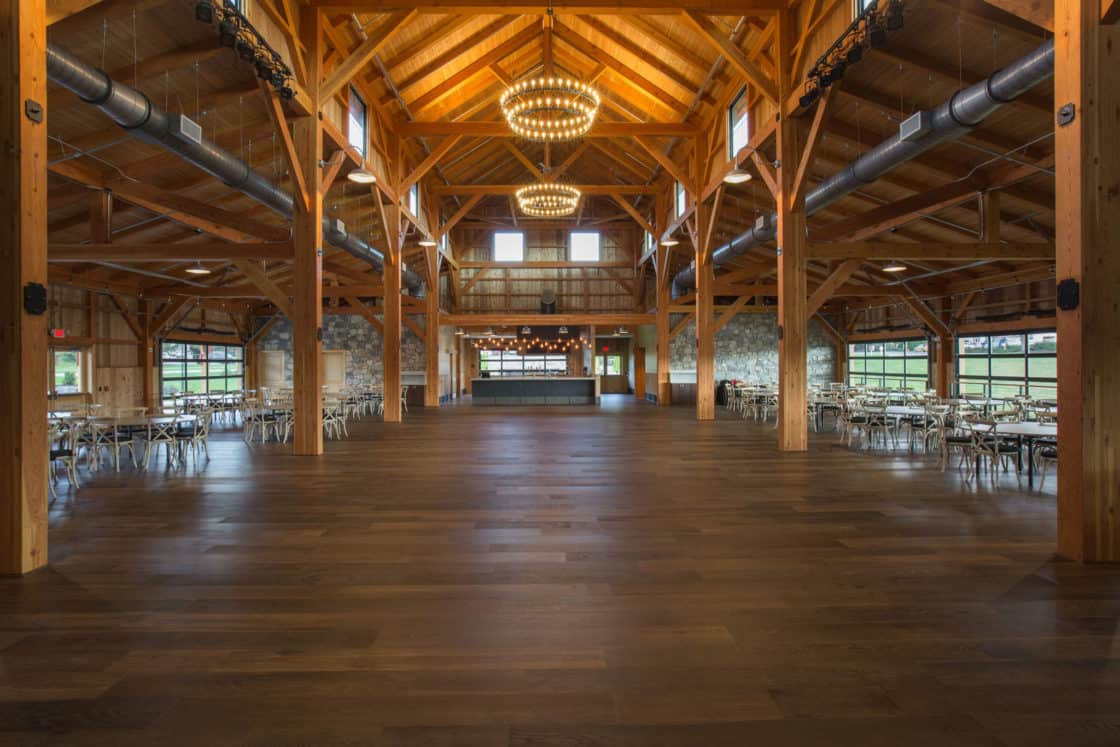 Spacious party barn-style event hall with exposed wooden beams, large windows, round chandeliers, and rows of tables and chairs arranged along the sides of the polished wooden floor.