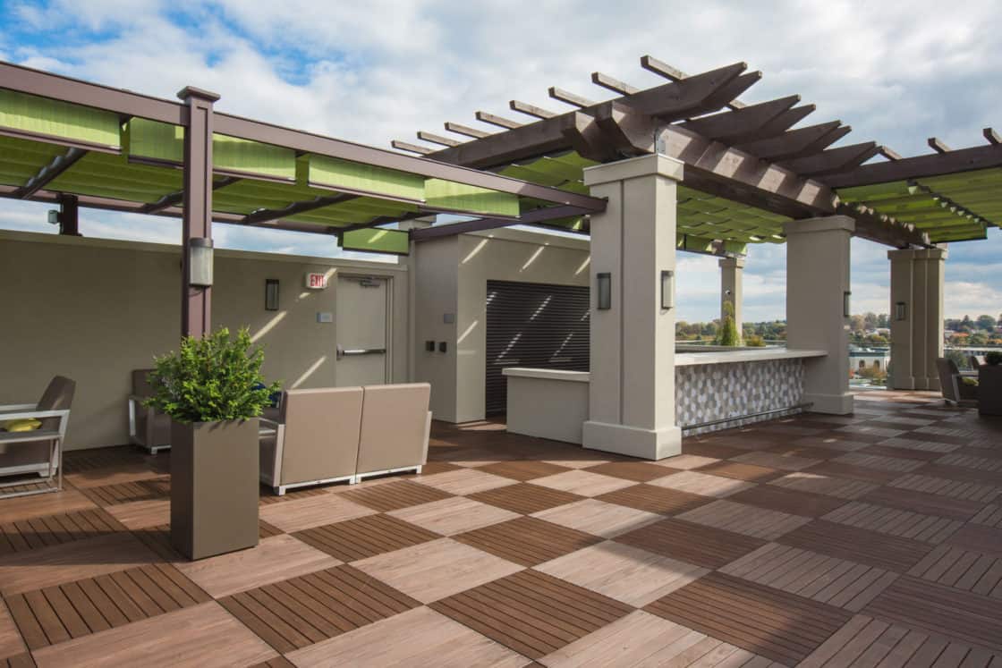A modern rooftop patio with wooden decking, pergolas with green shades, outdoor seating areas, and a bar counter—blending the relaxed elegance of a luxury home beneath a partly cloudy sky.