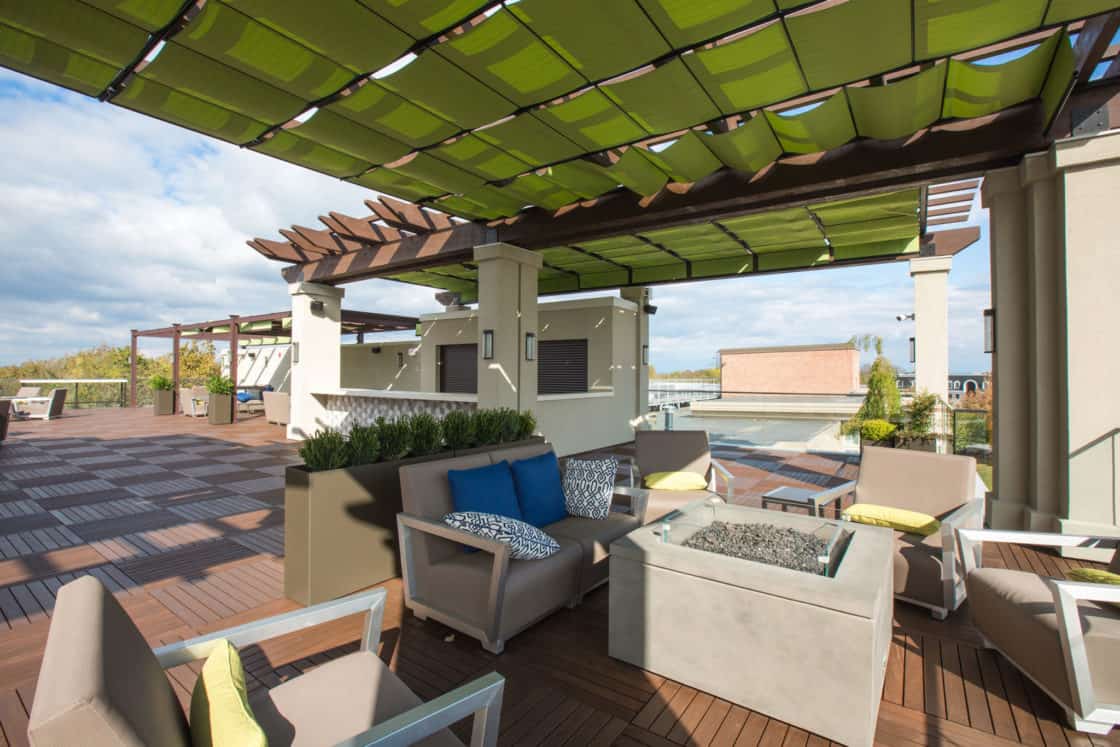 A spacious rooftop patio with wooden flooring, modern seating, potted plants, and a fire pit. Green shade panels are overhead, and buildings—reminiscent of a luxury home—are visible in the background under a partly cloudy sky.