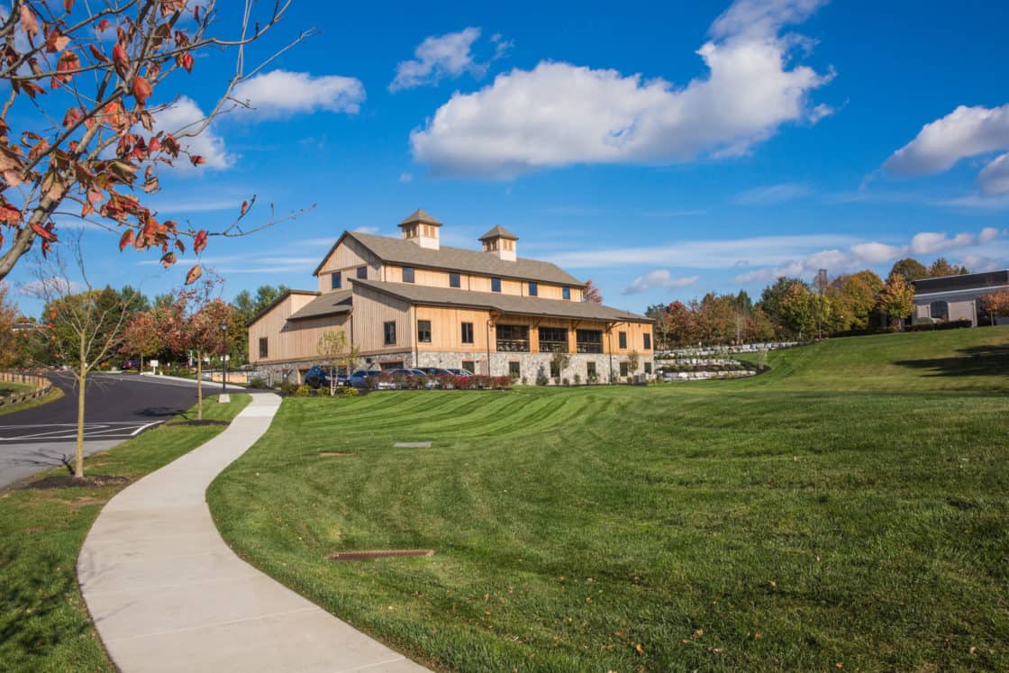 A large, modern bank barn-style building with stone and wood exterior sits on a manicured green lawn under a blue sky. A winding sidewalk leads toward this luxury horse barn, surrounded by autumn trees.