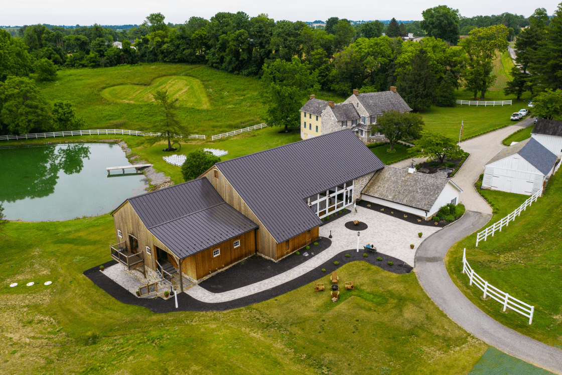 Aerial view of a rural property featuring a luxury horse barn, historic farmhouse, pond with dock, and winding driveway, all surrounded by green lawns, trees, and white fences.