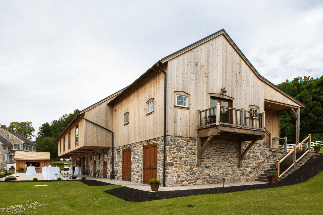 A large party barn with wood siding and a stone foundation, featuring a balcony, double doors, and stairs. Green grass surrounds a patio setup with white tablecloths, while trees provide a scenic backdrop.