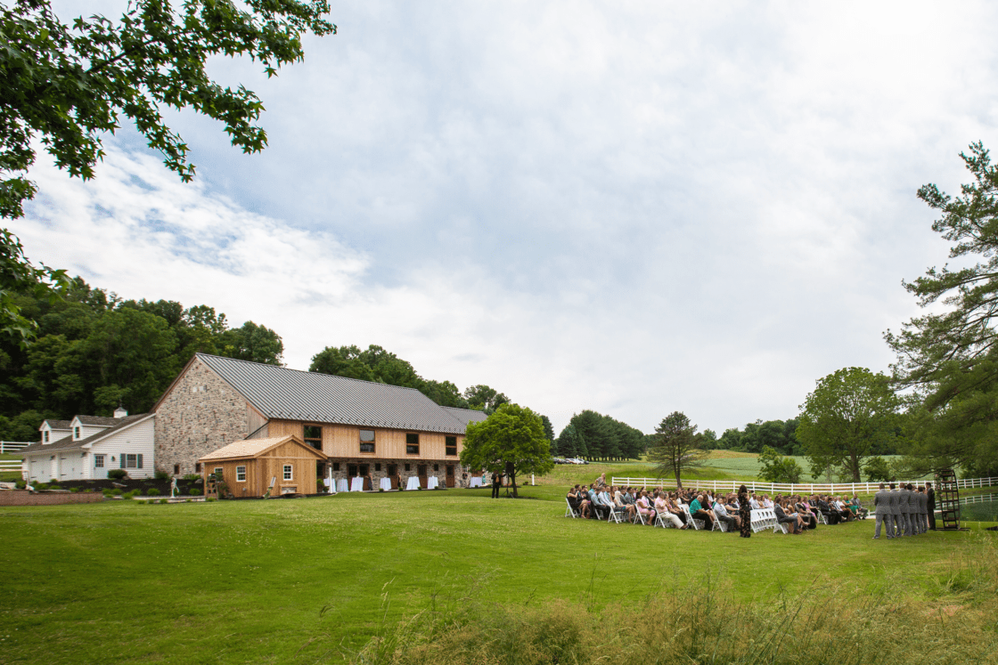 A wedding ceremony unfolds outdoors on a grassy lawn beside a historic bank barn, with guests seated in rows of white chairs. Trees and a white fence border the scenic, rural setting beneath a partly cloudy sky.