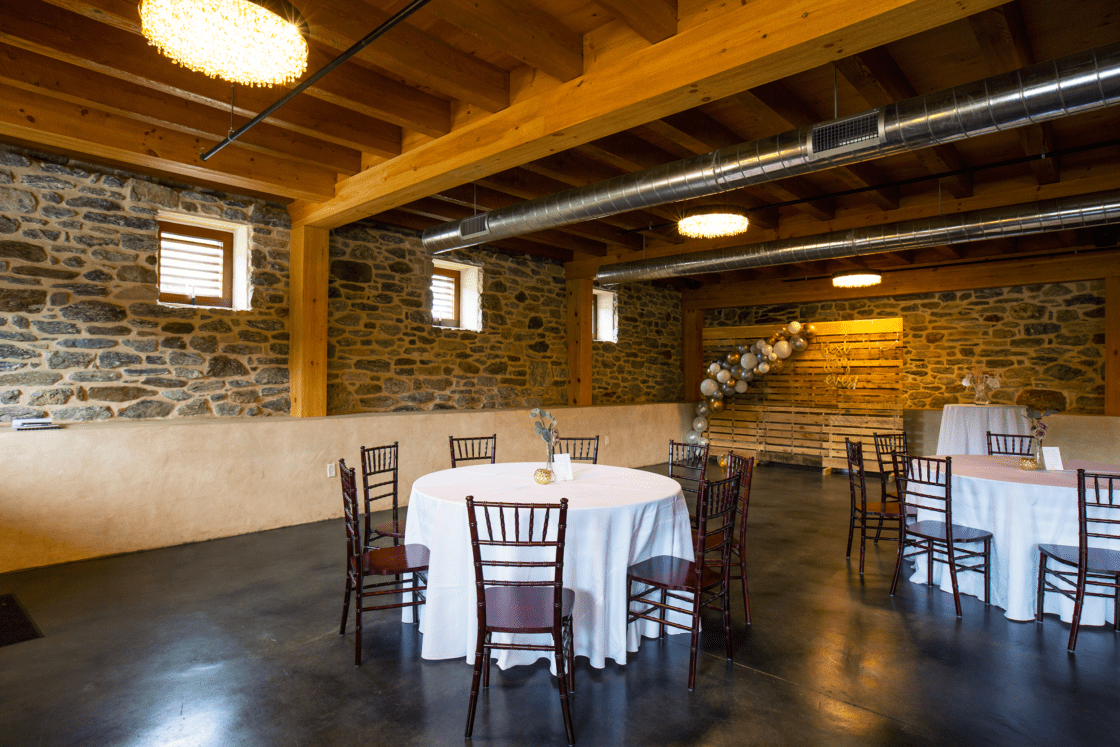 A rustic bank barn event space with stone walls, exposed wooden beams, and round tables draped in white cloths, each encircled by wooden chairs. Balloons decorate the corner, and the polished concrete floor adds charm.