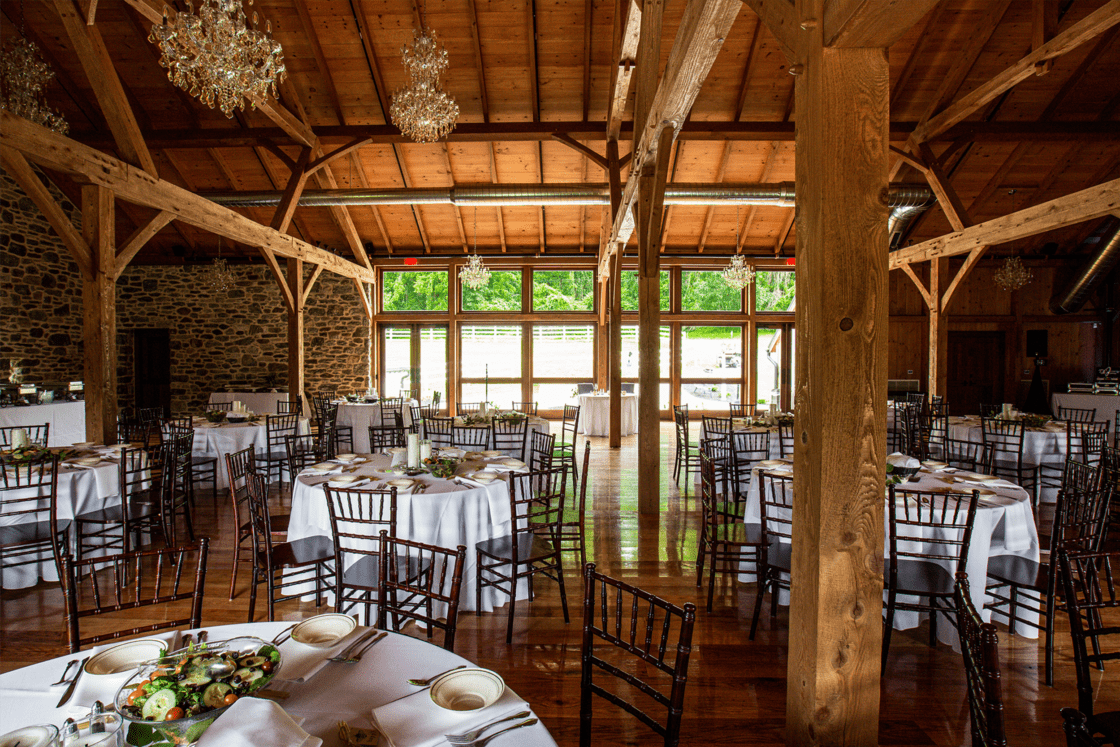 A rustic party barn with wooden beams and stone walls, set up for an event with round tables covered in white tablecloths, elegant place settings, and wooden chairs. Large windows fill the space with natural light.