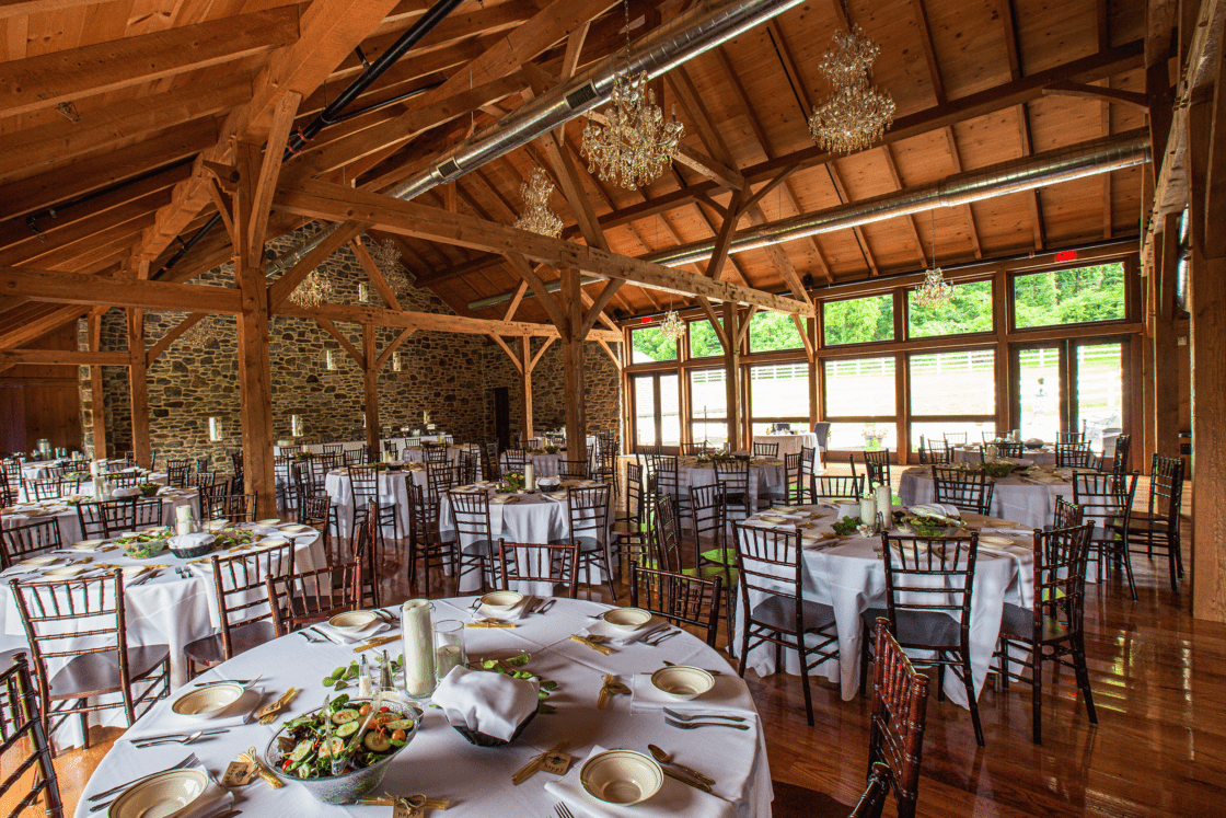 A rustic bank barn banquet hall with exposed wooden beams, stone walls, and large windows. Round tables with white tablecloths, dishes, and greenery centerpieces create an inviting atmosphere for an event or wedding reception.