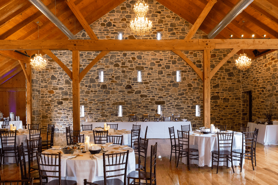 A rustic party barn with stone walls, exposed wooden beams, chandeliers, and round tables set with white tablecloths and dark wooden chairs, prepared for an event.