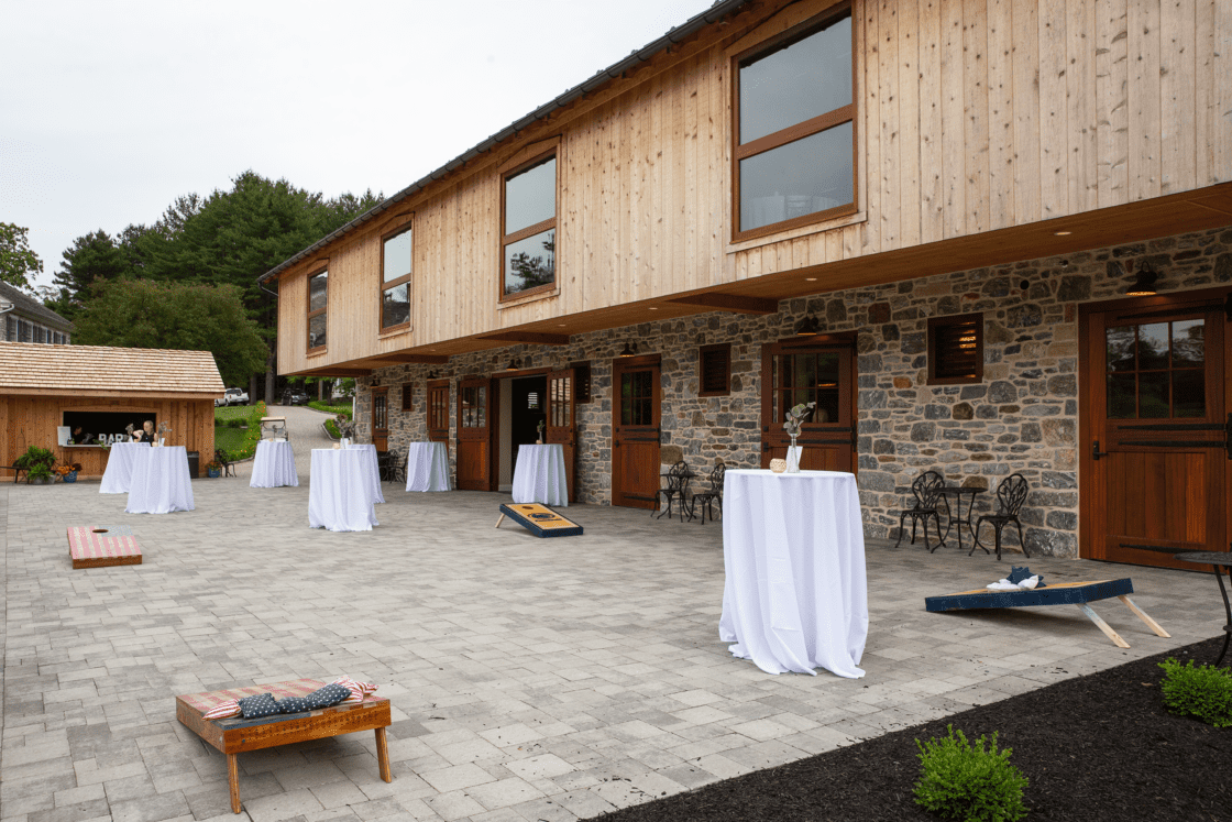 A spacious outdoor patio beside a luxury horse barn, with stone and wood accents, high tables draped in white cloths, cornhole boards on the paved ground, and lush greenery in the background—perfectly set up for an event.
