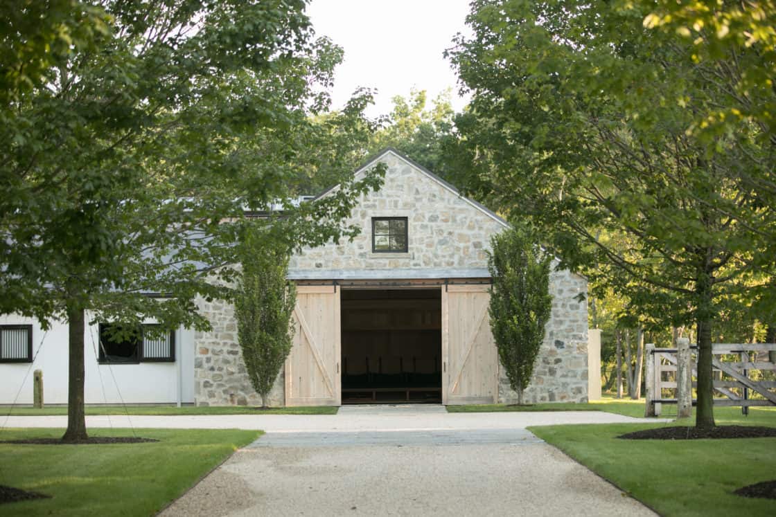 equine facility with open wooden doors sitting between green trees beneath blue sky