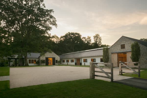 A modern farmhouse with stone buildings, metal roofs, and wooden doors sits behind a wooden gate, surrounded by trees and a gravel driveway at sunset—complete with a luxury horse barn for elegant country living.