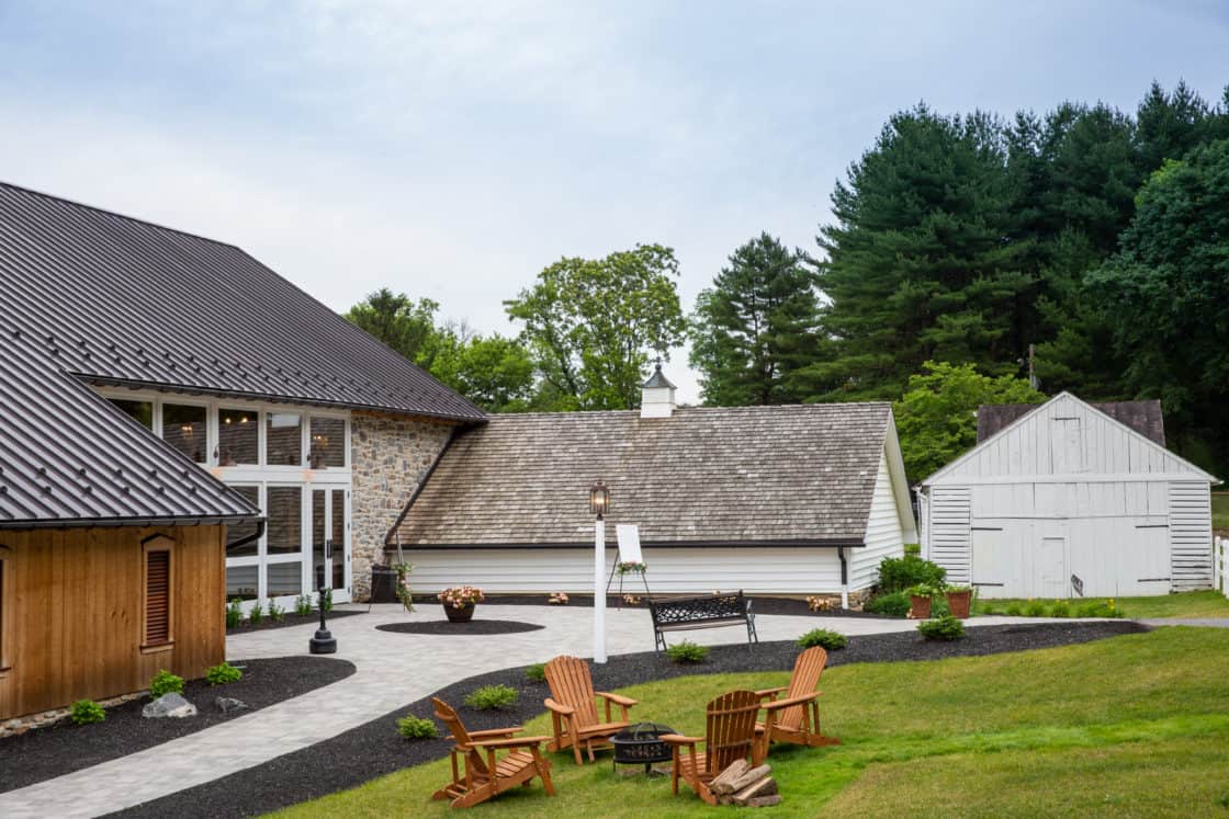 A landscaped courtyard features Adirondack chairs around a fire pit, with a modern party barn, a smaller rustic building, and a white shed surrounded by trees and greenery in the background.