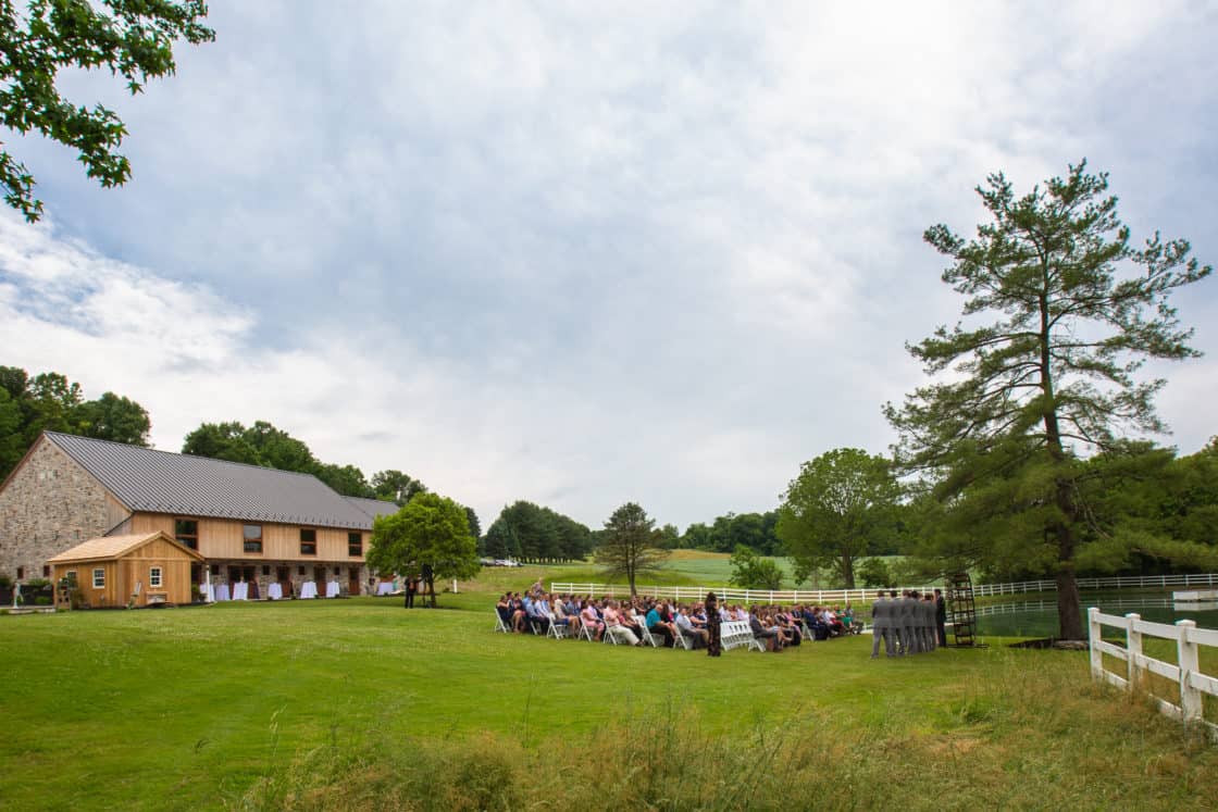 An outdoor wedding ceremony unfolds on a grassy lawn near a charming bank barn, with guests seated facing an arch. Tall trees and white fences frame the scenic countryside setting beneath a partly cloudy sky.