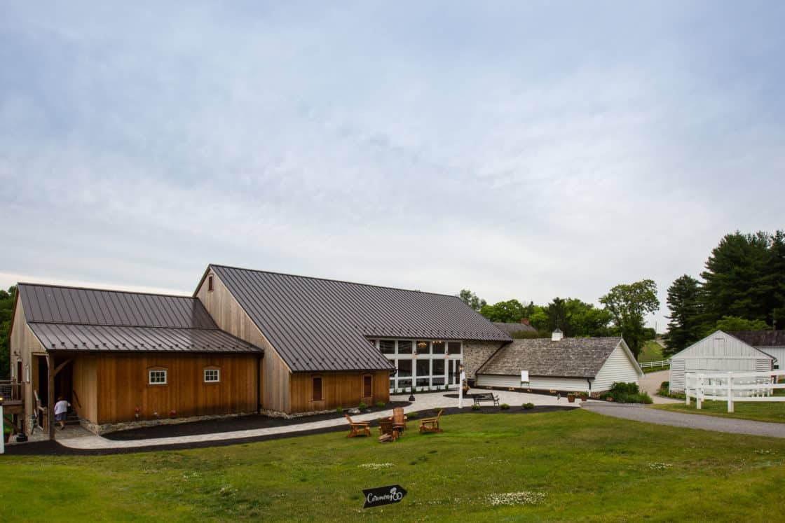 A large wooden bank barn with a metal roof sits on a green lawn, surrounded by trees and white fences under a cloudy sky. Wooden chairs are arranged outside, and a person stands near the barn entrance.