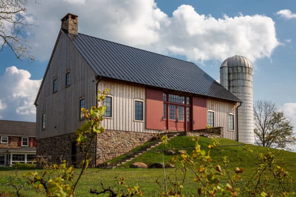 A large bank barn with a stone foundation, wooden siding, and a metal roof sits on a grassy hill next to a tall, white silo under a partly cloudy sky. Shrubs and rocks are visible in the foreground.
