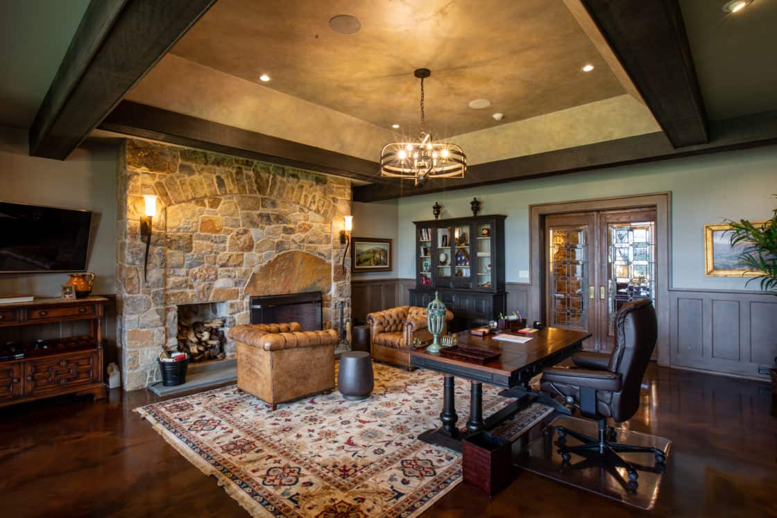 A luxury home office featuring a large stone fireplace, leather tufted sofa, wooden desk and beams evokes the charm of a bank barn. French doors and a wall-mounted TV complete this elegant retreat with bookshelves and warm lighting.