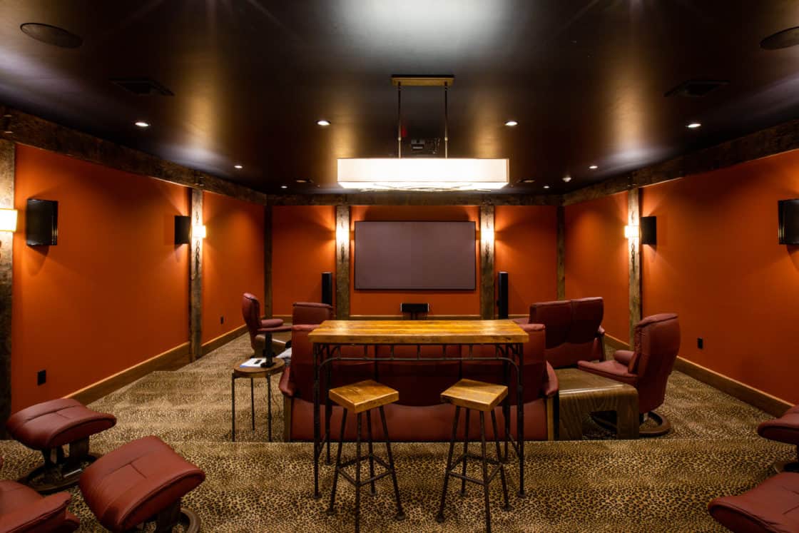 This luxury home theater room features orange walls, a large screen, and red recliners. A tall wooden table with stools sits at the center, while dim wall lights and a spotted carpet pattern create a cozy, inviting atmosphere.