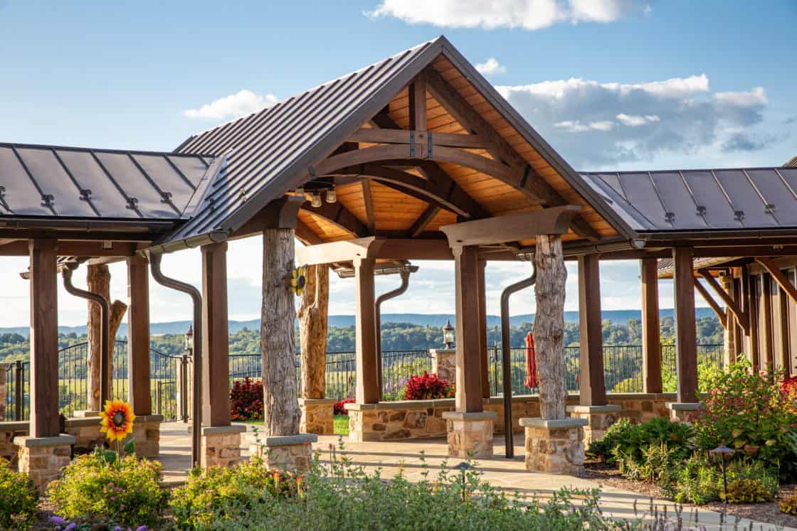 A wooden pavilion with a peaked roof and stone pillars stands in a landscaped garden, resembling a party barn, overlooking scenic green hills and a blue sky with scattered clouds.