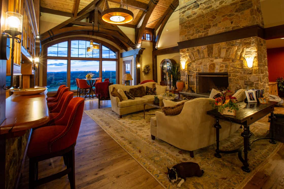 A cozy living room in a luxury home with a high wooden ceiling, stone fireplace, beige sofas, red bar stools, and a large window framing a scenic sunset. Warm lighting and a dog on the rug create an inviting, welcoming atmosphere.