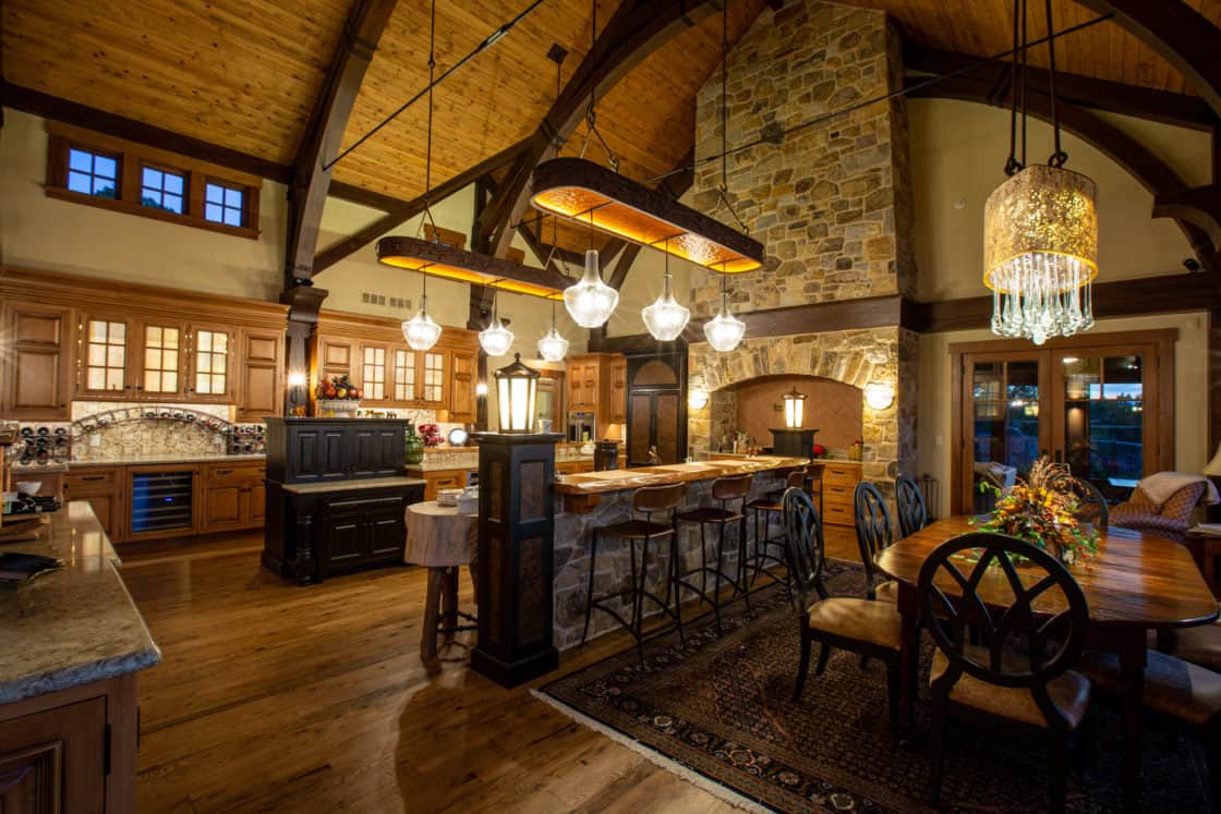 Spacious, rustic kitchen with exposed wood beams, stone accents, pendant lights, and a large island with barstools—this inviting space channels the charm of a bank barn with wooden cabinets, a dining table, and warm lighting for gatherings.