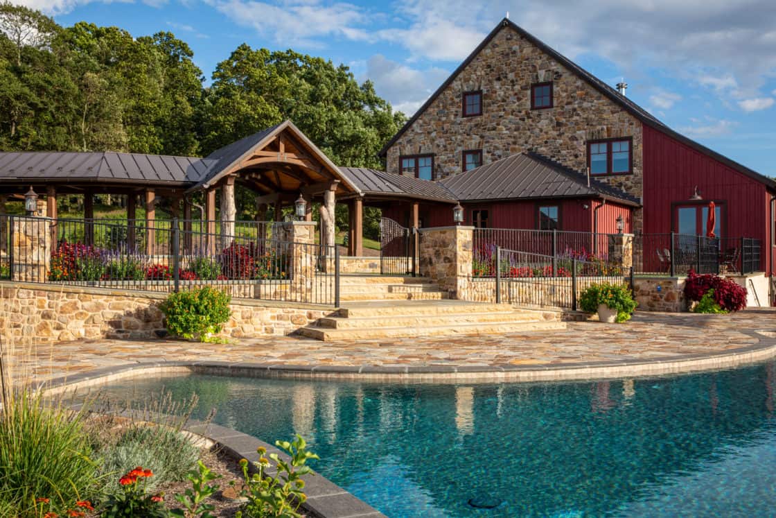 A luxury home with stone and wood details and red siding sits behind a landscaped patio and steps leading to a clear blue pool, surrounded by flowers and greenery, reminiscent of an elegant bank barn under a partly cloudy sky.