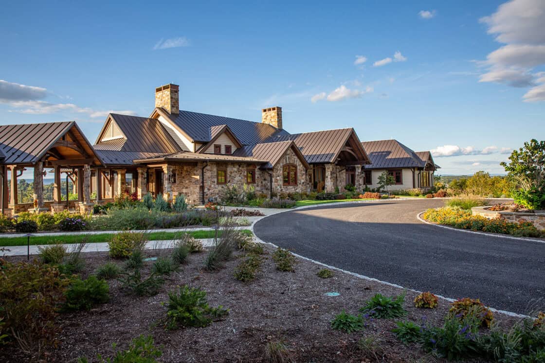 A large rustic stone house with a metal roof sits beside a luxury horse barn, surrounded by landscaped gardens and a circular driveway under a clear blue sky with scattered clouds.
