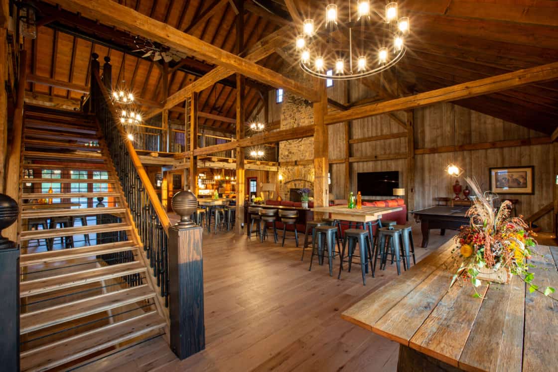 Spacious rustic interior with exposed wooden beams, large staircase, long wooden dining table, high ceilings, bar seating area, and chandeliers—this party barn’s warm lighting creates a cozy, inviting atmosphere perfect for gatherings.