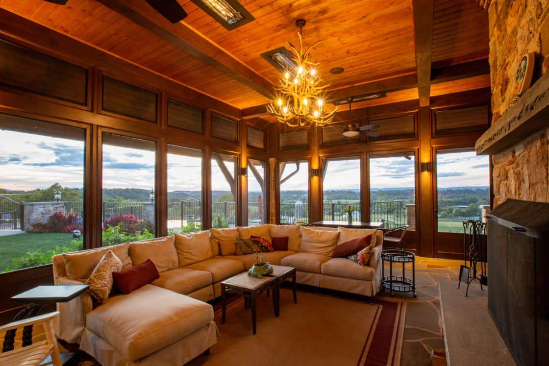 A cozy sunroom with a large beige sectional sofa, rustic wooden ceiling, chandelier lighting, and stone fireplace offers a scenic sunset view—perfect for relaxing after time spent at the nearby luxury horse barn.