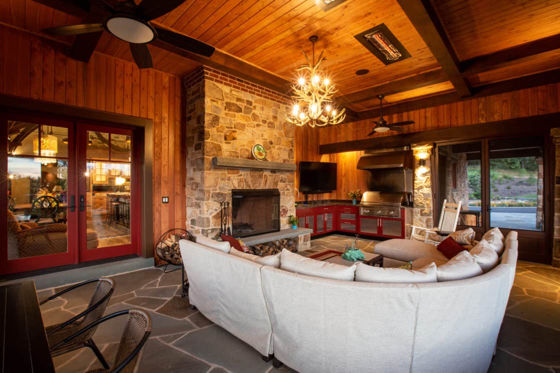 A cozy outdoor living space in a luxury home featuring a curved white sofa, stone fireplace, wood-paneled walls and ceiling, chandelier lighting, TV, and an outdoor kitchen with red cabinets and large windows.