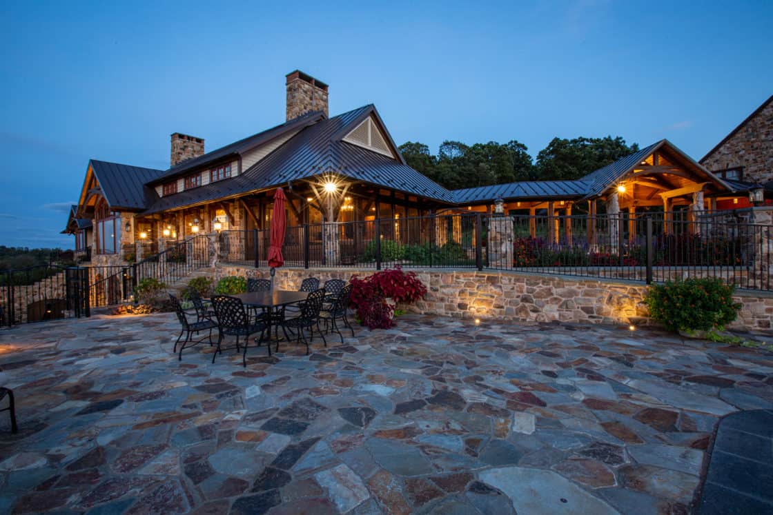 Rustic stone patio with wrought-iron tables and chairs sits before a large party barn with stone walls, wooden beams, and outdoor lighting, surrounded by trees at dusk.