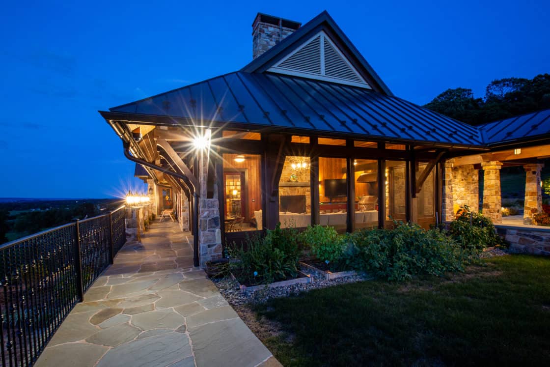 A warmly lit stone and wood luxury home with large windows and a metal roof at dusk, featuring a stone walkway, covered patio, and garden beds, surrounded by greenery and an expansive view.