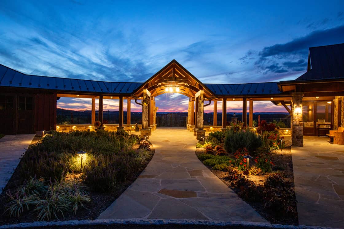 A warmly lit luxury home with a covered walkway and stone pillars is surrounded by landscaped gardens at dusk, set against a vibrant sunset and cloudy sky in the background.