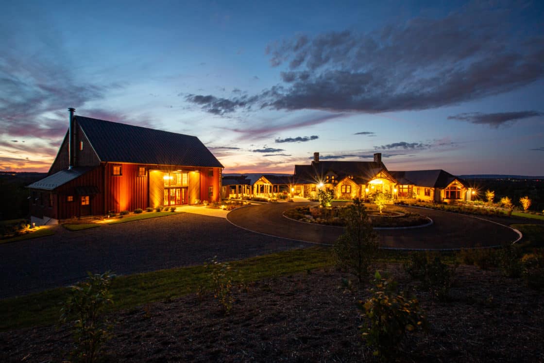 A warmly lit farmhouse and bank barn sit beside a circular driveway at dusk, with a dramatic blue and orange sky and scattered clouds in the background. Outdoor lights illuminate the rustic buildings and the surrounding landscape.