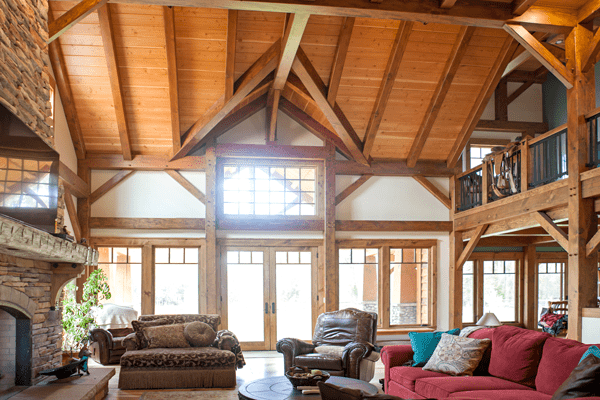 Spacious living room in this luxury home features a high vaulted wood ceiling with exposed beams, large windows, a stone fireplace, and cozy furniture including a red sofa. Natural light fills the room, creating an inviting retreat.