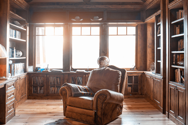 A cozy luxury home library with wooden bookshelves, a large leather armchair, books neatly arranged, and sunlight streaming through three large windows, creating a warm, inviting atmosphere.