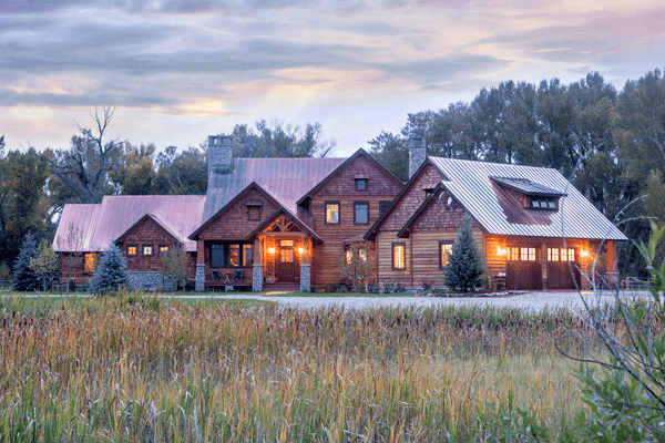 A large rustic wooden luxury home with warm lights glowing inside, surrounded by grass and tall trees at sunset, evokes the charm of a classic bank barn beneath a cloudy sky.