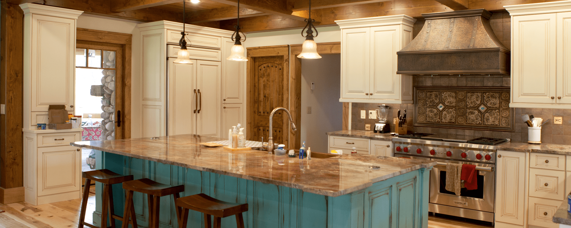 Rustic kitchen with wooden beams, cream cabinets, a large blue island with marble countertop—perfect for a luxury home or party barn—three wooden stools, pendant lights, and a stove with decorative tile backsplash and metal range hood.