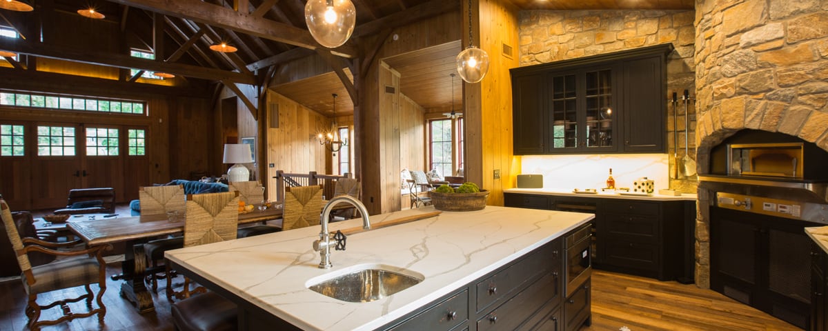 A rustic kitchen with a marble island, farmhouse sink, black cabinets, stone walls, and wood beams exudes charm in this luxury home. Warm lighting and an open dining and living area with wooden furniture evoke the cozy feel of a classic bank barn.