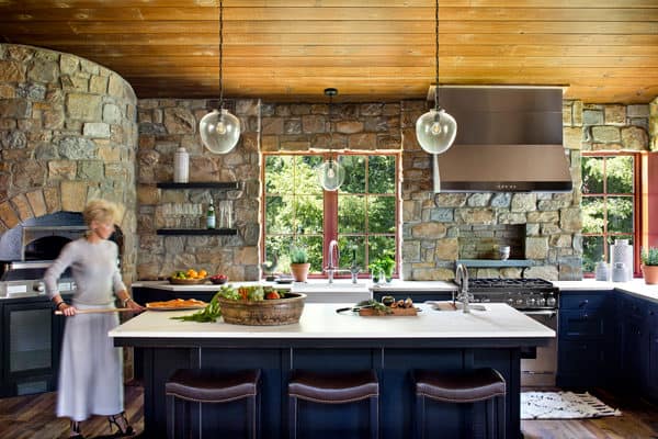 A rustic kitchen with stone walls, wooden ceiling, and large windows exudes the charm of a luxury home. A woman stands by the counter, surrounded by food and baskets. Modern appliances, open shelves, and dark cabinetry complete the inviting space.